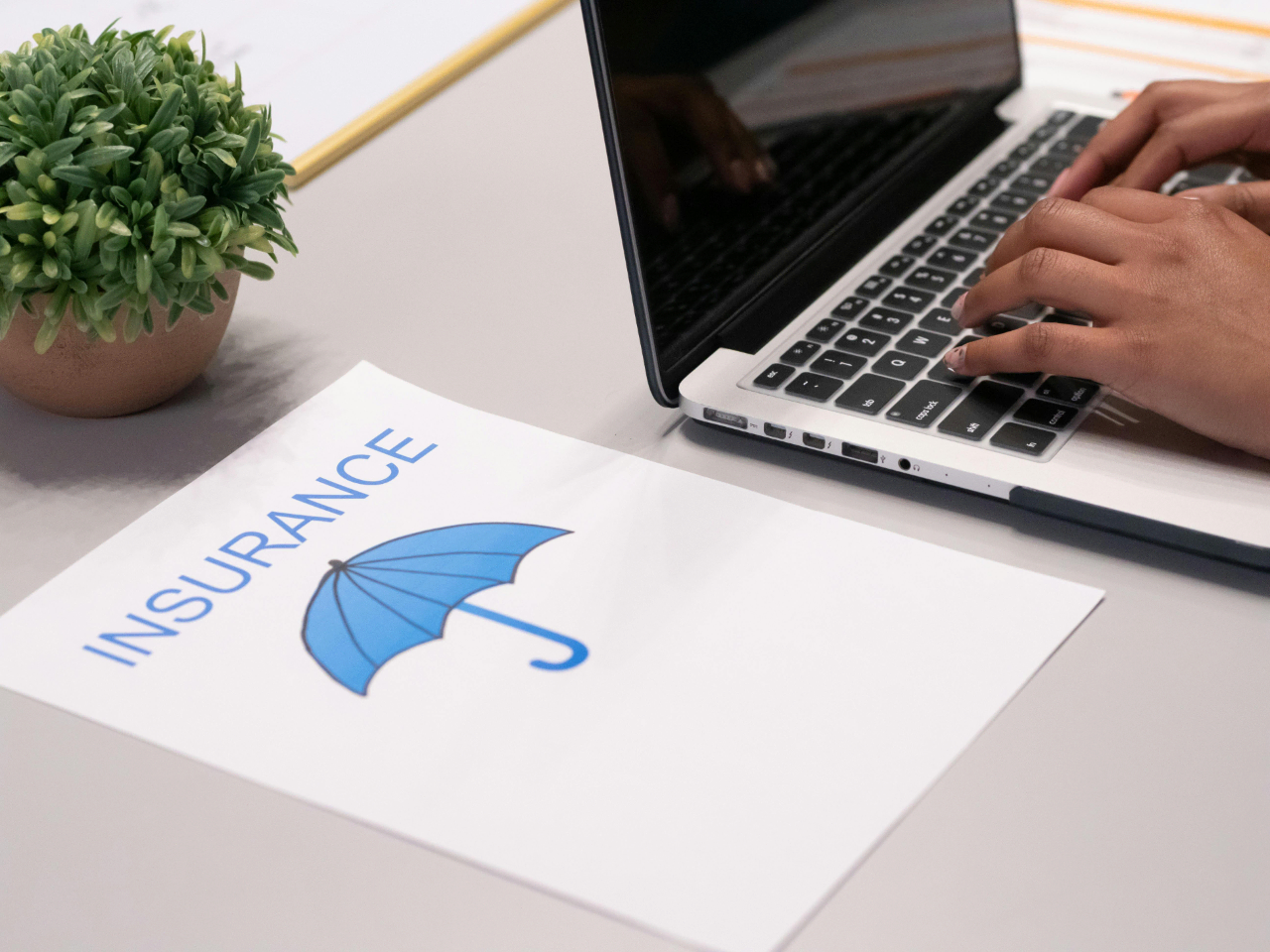 A piece of paper that reads "INSURANCE" and has a picture of an embrella on it sits on a table. To its right, someone is typing on a laptop and a plant is placed on the table above it.